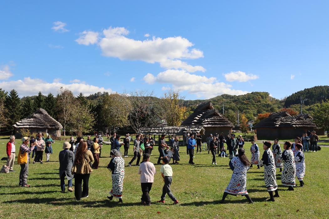 チセの前で皆が輪になりアイヌ古式舞踊を踊る写真