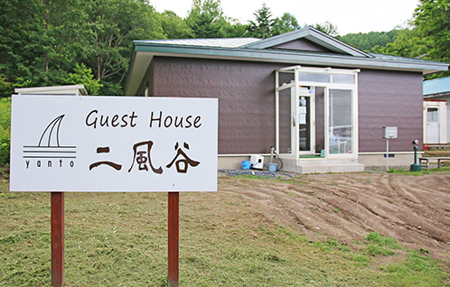 Guest House 二風谷 yantoと書かれた白色の看板の後ろに茶色を基調とした外壁に緑の屋根のゲストハウス二風谷ヤントの外観写真
