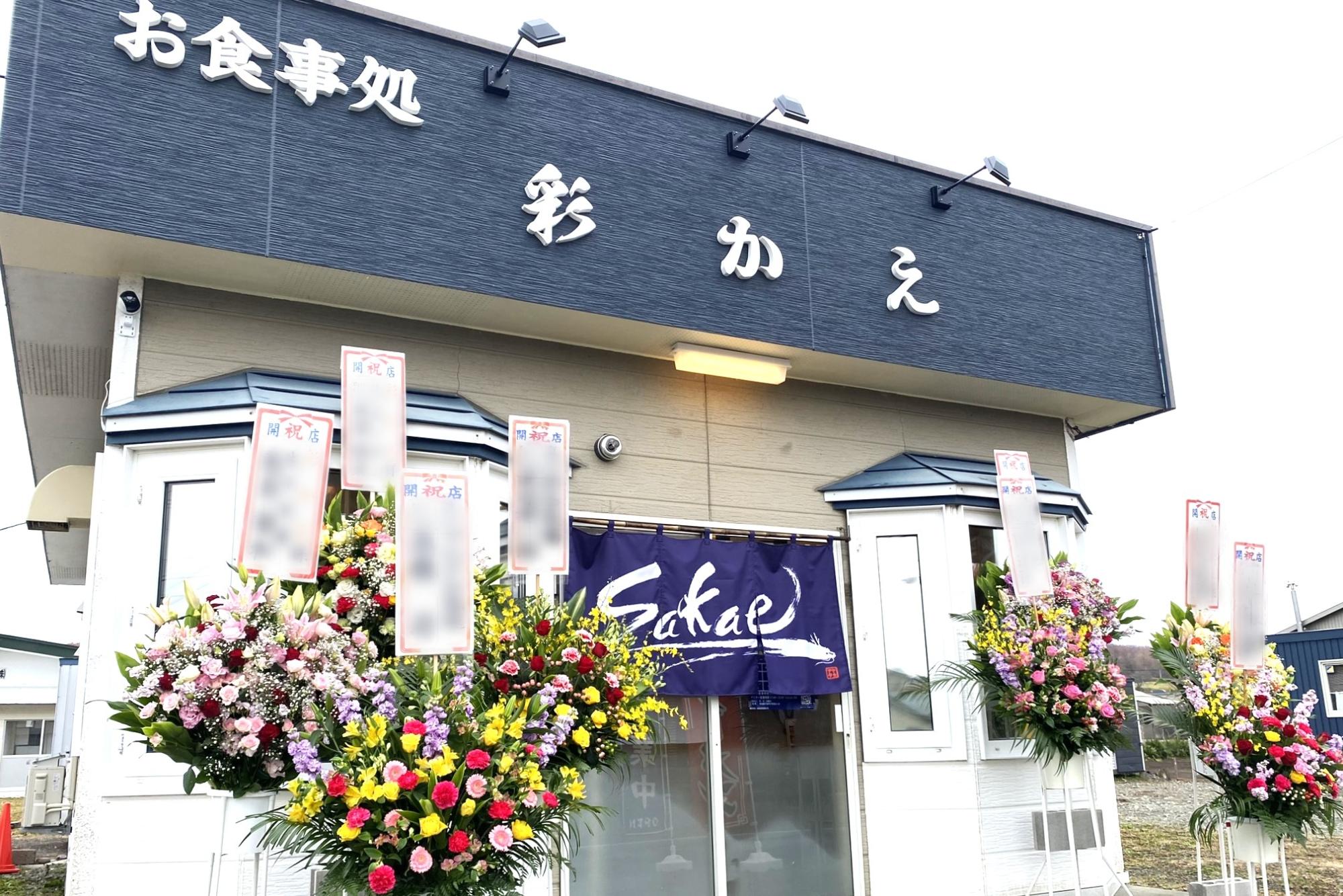 お食事処彩かえと書かれた建物の前に開店祝いの花が飾られている写真