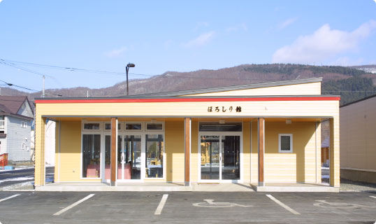 「ほろしり館」と書かれた黄色の建物で、建物の前には駐車場もある山の駅ほろしり館の外観写真