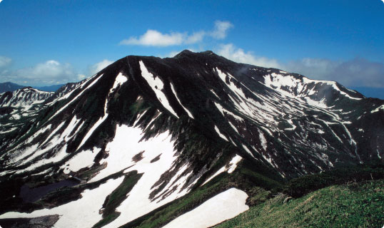 青空のもと、画面いっぱいに山々が連なっており、ところどころ雪で白くなっている幌尻岳の写真