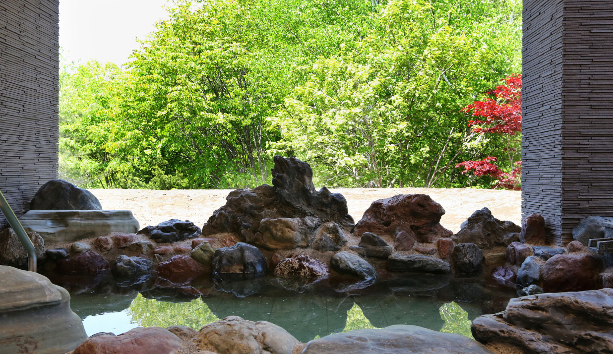 若草色の木々や赤く染まった紅葉を背景に大小の銘石を配した岩で造られた開放感あふれる露天風呂の写真