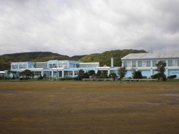 手前に広いグラウンドが広がっており、その奥に白と青を基調とした2階建ての建物の紫雲古津小学校の外観写真