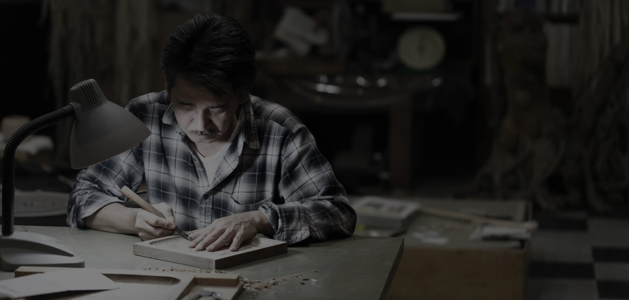 手元だけ照らされたうす暗い作業場で、真剣な表情で木彫り作業をしている貝澤 守さんの写真