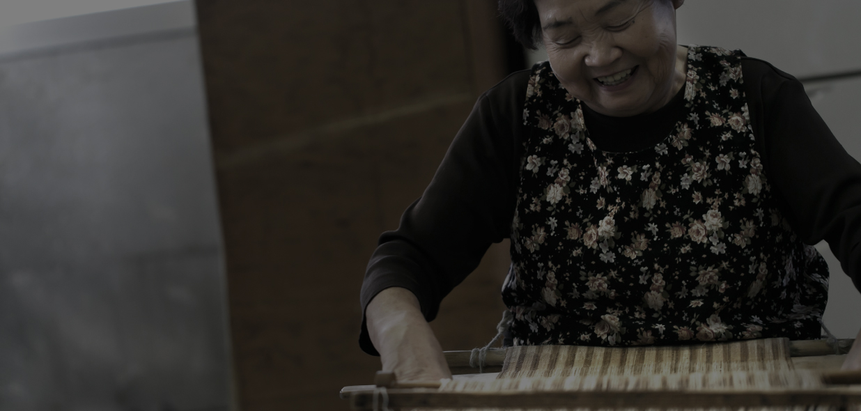 エプロンを身に着け、織り機の前で優しい笑みを浮かべながらでアットゥシ織りをしている貝澤 雪子さんの写真