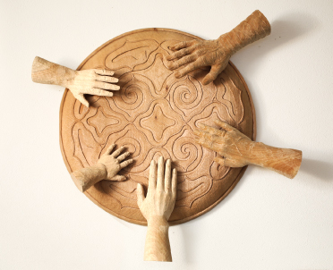 渦や柔らかい線で描かれた模様が入った丸い円盤に肌色の濃さや大きさが違う5つの手が添えられている貝澤 徹さんの木彫りの代表作品「ウコウク」の写真