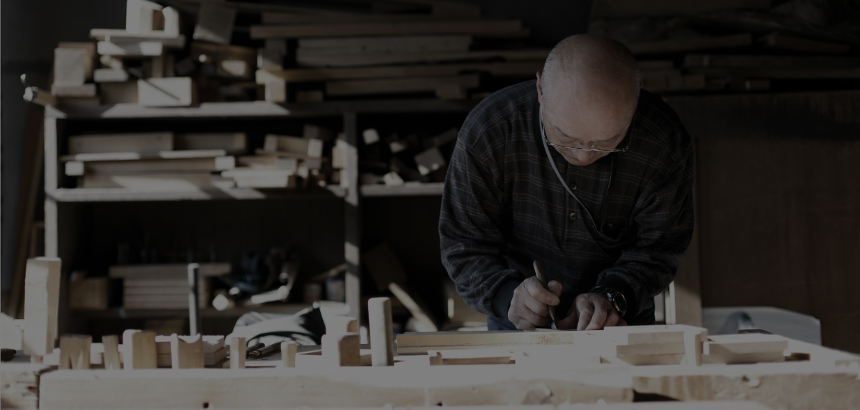 棚に大量の木材が置かれその手前の卓上で眼鏡をかけ慎重な手つきで手元を見ながら木彫り作業をしている洲崎 春男さんの写真